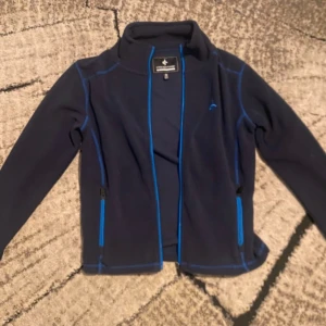 Blå fleecejacka från Cross Sportswear - Mörkblå fleecejacka från Cross Sportswear med blå detaljer och dragkedjor. Jackan har lång ärm, hög krage och två fickor med dragkedja. Perfekt för kyliga dagar och outdoor-aktiviteter. Snygg broderad logga på bröstet och kontrastsömmar.