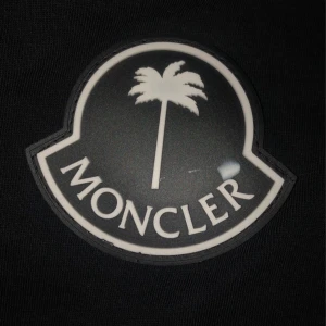 Svart Moncler t-shirt med palmtryck - Svart t-shirt från Moncler i storlek XL. Snyggt palmtryck på bröstet och en liten palm-detalj på ärmen. Klassisk rund hals och korta ärmar. T-shirten är gjord i mjuk bomull och har en clean, stilren vibe som passar perfekt till jeans eller shorts.