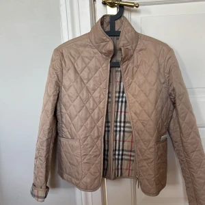 Beige quiltad jacka från Burberry,strl S - Snygg beige quiltad jacka från Burberry med klassiskt rutigt foder och guldfärgade knappar. Jackan har ståkrage, två fickor och dragkedja framtill. 