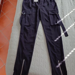 Svarta cargopants från Bik Bok, strl 36 - Snygga svarta cargopants från Bik Bok med stora fickor på sidorna, dekorativa ringar och långa band. Dragkedjor vid bensluten ger en cool touch. Byxorna har normal passform och är tillverkade i ett mjukt bomullsmaterial. Perfekta för dig som gillar streetstyle.