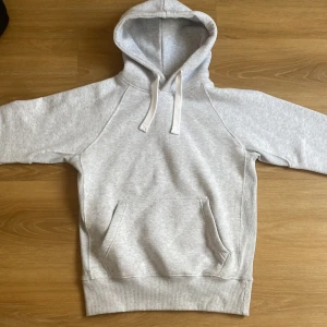Russemerch Ljusgrå hoodie med magficka - Ljusgrå hoodie med klassisk magficka och huva med vita snören. Tröjan har långa ärmar och ribbade muddar vid ärmslut och nederkant. Perfekt för chill dagar eller när du vill ha en enkel och stilren look.