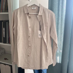 Beige linneskjorta från Kappahl - Snygg beige skjorta från Kappahl i storlek 152. Skjortan är tillverkad i linne, har klassisk krage, långa ärmar och knäppning framtill. Perfekt för en clean och stilren look. En bröstficka ger extra detalj. Helt ny