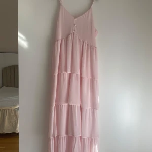 Ljusrosa långklänning med volanger - Supersöt ljusrosa långklänning med flera volanglager och smala axelband. Klänningen har knappar framtill och smock i ryggen. Använd 1 gång. 