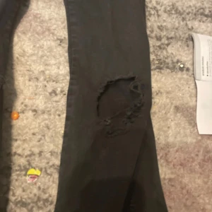 Svarta bootcut jeans med hål - Säljer ett par svarta jeans med bootcut passform och stora slitna hål vid knäna. Jeansen har klassisk femficksdesign och är gjorda i jeansmaterial med lite stretch. Perfekt för dig som gillar en edgy och avslappnad stil.