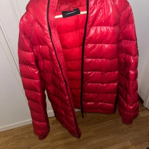 Peak Performance Dunjacka - Tja! säljer en röd dunjacka från Peak Performance med huva och svarta detaljer. Jackan har quiltad design, dragkedja framtill och logotyp på ärmen. Perfekt för kalla dagar och har en snygg, sportig look som sticker ut. Sticker även ut med sin glans.