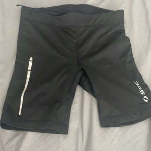 Svarta träningsshorts från SOC - Svarta träningsshorts från SOC med midwaist passform. Snyggt sportig look med snörning i midjan och praktisk dragkedja på bakfickan. Materialet är syntet och designen är tight, perfekt för gym eller löpning.
