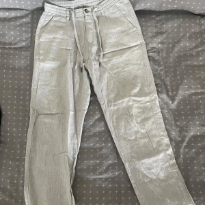 Ljusgrå linnebyxor från Lindbergh - Ljusgrå linnebyxor från Lindbergh med dragsko i midjan och klassiska fickor fram och bak. Byxorna är tillverkade i en skön mix av bomull och linne, perfekta för varmare dagar. Stilsäker och avslappnad look med raka ben och bälteshällor.