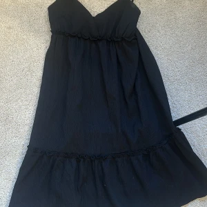 Svart klänning från H&M S - Svart kort klänning från H&M Divided med smala axelband och v-ringad omlott upptill. Klänningen har volangdetalj nertill och är tillverkad i ett lätt, krinklat tyg som ger en avslappnad känsla. Perfekt för dig som gillar enkel men snygg stil.