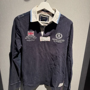 Marinblå pikétröja från Henri Lloyd - Snygg marinblå långärmad pikétröja från Henri Lloyd med broderade detaljer, brittisk flagga och vit krage med ljusblå inslag. Klassisk och sportig look med knappar i halsen och broderad logga på bröstet. Perfekt för dig som gillar preppy stil.