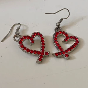 Silvriga örhängen med röda hjärtan - Coola silvriga örhängen i form av hjärtan dekorerade med röda glittrande stenar. Hängande modell med krok, ca 4 cm långa. Perfekta för att ge din look en extra pop av färg och kärlek.