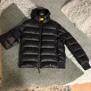 Svart dunjacka Parajumpers Sheen XL - Säljer en svart dunjacka från Parajumpers, modell Sheen, i storlek XL. Jackan är helt ny fick i somras men är tyvärr lite förliten nu. Jackan har en klassisk pufferstil med huva, gul detalj vid halsen och dragkedja framtill. Insidan är ljusgrå och jackan har ribbade muddar vid ärmslut och nederkant. Perfekt för kalla dagar! 
