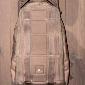 Beige ryggsäck från Db - Säljer en stilren beige ryggsäck från Db med rektangulär form och diskret logga framtill. Väskan har flera praktiska fack, robusta dragkedjor och vadderade axelremmar med metallspännen. Perfekt för skola eller cityhäng.