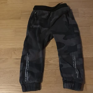 Svarta camo mjukisbyxor med dragkedjor - Snygga svarta mjukisbyxor med mörkgrått camouflagemönster och dragkedjor på fickorna. Resår i midjan med snörning och muddar vid bensluten. Perfekta för chill eller streetwear.