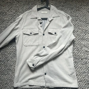Ljusgrå overshirt med knappar - Snygg ljusgrå overshirt från Jack & Jones med klassisk krage och svarta knappar. Två bröstfickor med lock och långa ärmar med knapp vid mudden. Perfekt att bära som lager över t-shirt eller tröja. Stilren och enkel design. Väldigt bra skick endast lite skrynklig. Storlek xl men sitter som L