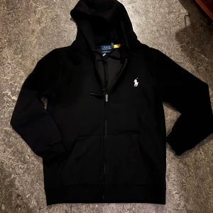 Svart hoodie från Polo Ralph Lauren - Svart hoodie från Polo Ralph Lauren med vit broderad logga på bröstet. Tröjan har dragkedja, snörning vid huvan och två fickor framtill. Slim fit-modell som sitter snyggt och har långa ärmar. Perfekt för chill dagar eller när du vill ha en clean look.