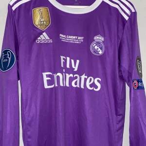 Real Madrid Ronaldo lila långärmad tröja - Säljer en officiell Real Madrid Champions League Final Cardiff 2017 tröja med Ronaldo 7 på ryggen. Långärmad, lila med vita detaljer och broderade märken. Adidas Aeroready-material för skön känsla. Fly Emirates tryck på bröstet och Real Madrid-logga. 