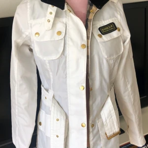Vit Barbour International jacka  - Snygg vit jacka  från Barbour International med guldiga knappar, svart krage i manchester och klassiskt rutigt innerfoder. Jackan har flera fickor, bälte i midjan och dragkedja framtill. Perfekt för dig som vill ha en stilren och sportig look. Storlek 34 men passar även 36 . Använd max 3 ggr pga för tajt för mig 