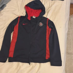 PSG x Nike marinblå hoodie XL - Säljer en fett snygg Paris Saint-Germain hoodie från Nike i marinblått med röda detaljer. Tröjan har huva, dragkedja och PSG-logga på bröstet. Perfekt för dig som älskar fotboll och vill ha en sportig stil. Passar dig som är 158-170 cm lång.