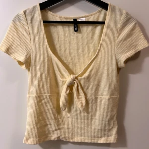 Ribbad ljusgul knyt-topp från H&M - Superfin ljusgul ribbad topp från H&M med korta ärmar och snygg knytning framtill. Toppen har en djup v-ringning och är i mjuk bomullsmix, perfekt för en chill och trendig look.