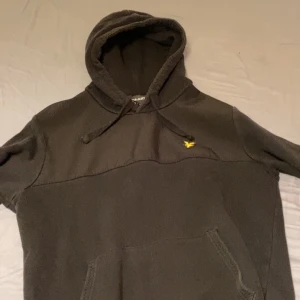 Svart hoodie från Lyle & Scott - Svart hoodie från Lyle & Scott med klassisk huva och snörning. Tröjan har en gul broderad logga på bröstet och en stor magficka framtill. Perfekt för dig som gillar en enkel och stilren look.