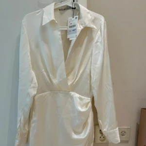 Ny Vit omlottklänning från Zara i satin - Snygg vit kortklänning från Zara i glansigt satinmaterial. Klänningen har lång ärm, klassisk krage och djup v-ringning med omlott-detalj framtill. Perfekt för dig som gillar stilrena och trendiga plagg med en lyxig känsla. Oanvänd 
