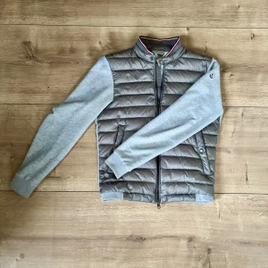 Grå cardigan från Moncler - Snygg grå cardigan från Moncler med quiltad framsida och mjuka, ljusgrå ärmar i bomull. Kragen har en cool randig detalj i rött, vitt och blått. Västen har dragkedja framtill och Moncler-logga på ärmen. Perfekt för lager-på-lager och streetstyle. Obs jackan har en liten gräs fläck på axeln men inget som är synligt om det inte är noga inspektion