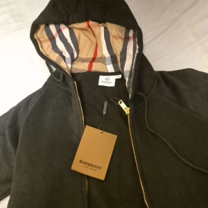 Svart burberry hoodie med dragkedja - Säljer en svart hoodie från Burberry med klassiskt rutigt foder i huvan och guldfärgad dragkedja. Tröjan har huva med snörning och är långärmad. Perfekt för dig som vill ha en stilren och lyxig look med ikonisk Burberry-touch.