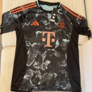 FC Bayern München Adidas fotbollströja - Snygg FC Bayern München fotbollströja från Adidas med svart bas och marmorerat mönster i ljusblått. Tröjan har korta ärmar, orange detaljer och klubbmärke på bröstet. Tillverkad i lätt och ventilerande material med Aeroready-teknologi.