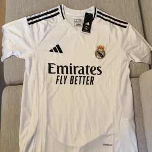Real Madrid hemmatröja Adidas vit - Snygg Real Madrid hemmatröja från Adidas i vit med svarta detaljer och klubbmärke på bröstet. Tröjan har korta ärmar, klassiska svarta ränder på axlarna och är tillverkad i lätt och ventilerande material. Perfekt för dig som älskar fotboll och vill visa ditt stöd för laget.