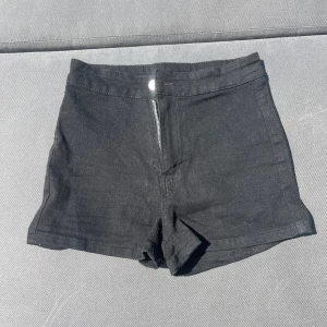 Svarta högmidjade jeansshorts - Svarta, klassiska högmidjade jeansshorts med dragkedja och knapp framtill. Shortsen har två bakfickor och en enkel, stilren design som passar till allt. Perfekta för varma dagar när du vill ha en clean och snygg look.