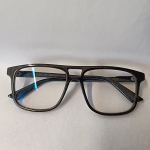 Svarta glasögon från Gucci - Snygga svarta glasögon från Gucci med markerad, fyrkantig båge i plast. Modellen är GG1035O 001 5516 och har en modern, stilren design med diskret logga på insidan av skalmen. Perfekta för dig som vill ha en klassisk men trendig look. Har styrka.