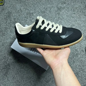 Maison Margiela  gats svarta sneakers - Maison Margiela  sneakers i svart skinn och mocka med vita snören och beige gummisula. Klassisk low-top siluett med rund tå och stilren design. Perfekta för dig som gillar minimalistisk och tidlös streetstyle.  Icke äkta