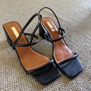 Svarta sandaler med blockklack - Stilrena svarta sandaler från Primark med fyrkantig tå och trendiga tunna remmar. Skorna har en blockklack på ca 7 cm och innersula i brunt syntetmaterial. Perfekta för dig som gillar minimalistisk och modern look.