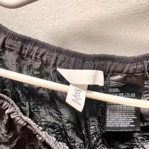 Svart långkjol från H&M i glansigt tyg - Svart långkjol från H&M med glansig, lätt skrynklig yta och resår i midjan. Kjolen har en enkel och stilren look som passar perfekt till sneakers eller boots. Materialet är en mix av polyester och polyamid för en lätt och luftig känsla.