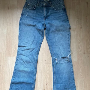 Blå bootcut jeans med hål på knät - Säljer ett par klassiska blå jeans med bootcut-snitt och ett stort slitet hål på höger knä. Jeansen har fem fickor, normal passform och är tillverkade i robust denim. Perfekta för en avslappnad och trendig look.