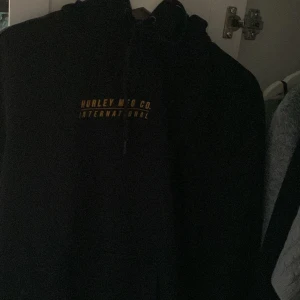 Svart hoodie från Hurley - Svart hoodie från Hurley med gul text på bröstet där det står 'Hurley MFG CO. International'. Klassisk modell med huva och snörning, perfekt för en chill och avslappnad stil.