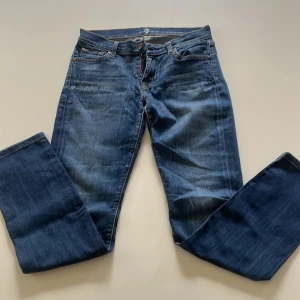 Blå jeans med broderade fickor - Snygga blå jeans från 7 For All Mankind med klassisk femficksdesign och gul broderad detalj på bakfickorna. Jeansen har låg midja och en rak passform som sitter tight upptill och släpper vid benen. Tillverkade i mjukt denimtyg med lätt tvättad look.