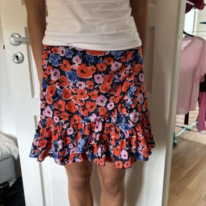 Blommig volangkjol från Gina Tricot - Supersöt kort kjol från Gina Tricot med färgglatt blommönster i rött, rosa och blått. Kjolen har volangkant nertill och en omlottliknande detalj framtill. Perfekt för dig som vill sticka ut med en lekfull och somrig vibe.