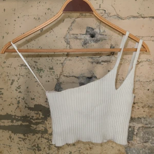 Vit ribbad croppad linnetopp - Snygg vit croppad linnetopp med ribbad struktur och asymmetriska axelband. Perfekt för varma dagar eller lager på lager. Toppens material är mjukt och stretchigt, vilket gör den bekväm och lätt att styla till olika outfits.