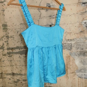 Blå linnetopp med rosett i satin - Supersöt blå linnetopp i glansigt satinliknande material med breda axelband och en stor rosett bak. Toppen har en luftig, lätt utställd passform och är perfekt för varma dagar. Färgen är klarblå och tyget har en fin lyster som verkligen poppar.