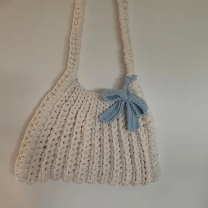 Virkad vit axelväska - Superfin handgjord axelväska i vitt. Väskan har en bred axelrem och en mjuk, chunky känsla som ger en trendig och mysig look. Rosetten ingår inte men går att köpa till för 50kr