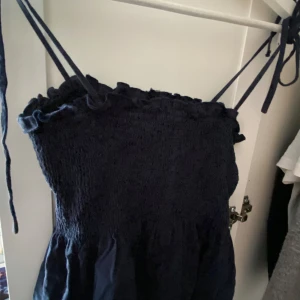 Mörkblå sommarklänning med smock - Supersöt mörkblå klänning med smockad överdel och tunna axelband som knyts. Volangdetalj upptill ger en gullig touch. Perfekt för varma dagar och enkel att styla med sneakers eller sandaler.