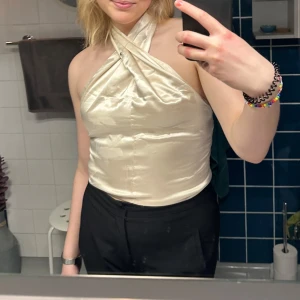 Champagnefärgad halterneck topp i satin - Snygg halterneck topp i champagnefärgad satin med omlott-detalj framtill och smockad rygg. Toppen är ärmlös och har en glansig finish som ger en lyxig känsla. Perfekt att styla med jeans eller kjol för en trendig look.