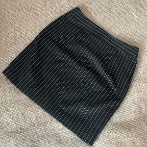 Svart kortkjol med vita ränder/ kostym kjol - Snygg svart kortkjol med kostym material. Kjolen har en rak passform och är tight.