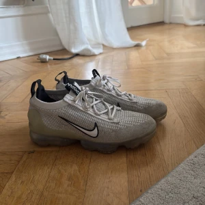 Nike Vapormax Flyknit vita sneakers - Nike Vapormax Flyknit sneakers i ljusgrått och vitt med svart swoosh. Ovandel i stickad flyknit för en luftig känsla och cool look. Genomskinlig sula med den klassiska Vapormax-dämpningen. Har dock ingen box till skorna.