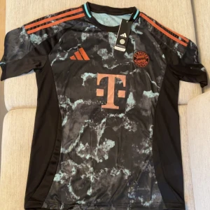 FC Bayern München Adidas fotbollströja - Snygg FC Bayern München fotbollströja från Adidas med svart och turkosmarmorerat mönster. Tröjan har korta ärmar, orange detaljer och klubbmärke på bröstet. Tillverkad i lätt funktionsmaterial med klassiska Adidas-ränder på axlarna.