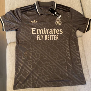 Real Madrid x Adidas grå fotbollströja - Snygg grå Real Madrid fotbollströja från Adidas med diskret mönster över hela tyget. Tröjan har korta ärmar, klassisk krage med vita detaljer och svarta Adidas-ränder på axlarna. Stort Emirates-tryck på bröstet och klubbmärke i vitt. Perfekt för dig som vill sticka ut på läktaren.