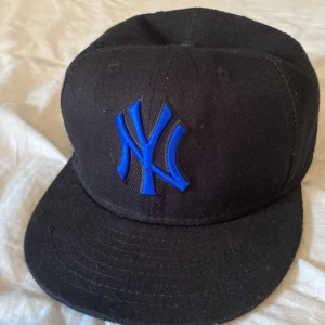 New York Yankees svart keps 7 1/2 - Svart keps från New York Yankees med blått broderat NY-logga framtill och blå MLB-logga bak. Klassisk rak skärm och stilren look, perfekt för dig som gillar sportig streetstyle. Tillverkad i bomull och passar dig som vill ha en ikonisk accessoar.