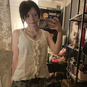 Vit broderad linnetopp från Asanga - Supersöt vit linnetopp från noa noa med broderade hålmönster och volangdetalj längs knäppningen framtill. Ärmlös och med v-ringning, perfekt för varma dagar. Lätt och luftig känsla tack vare bomullsmaterialet.