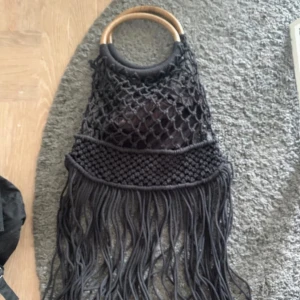 Svart handväska med fransar och trähandtag - Säljer en svart handväska med snyggt nätmönster och långa fransar. Väskan har runda trähandtag som ger en cool boho-vibe. Perfekt accessoar för att lyfta din outfit med en unik touch.